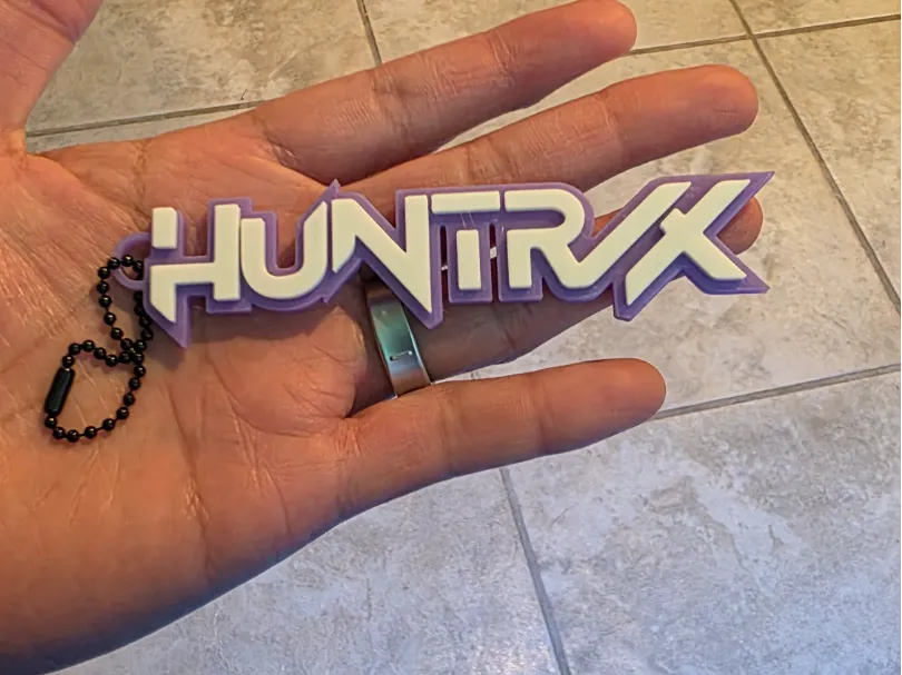 Purple and white 3D printed HUNTRIX logo keychain held in a hand